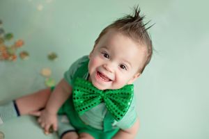 Baby wearing green bowtie smiles joyfully against mint green background during portrait session.