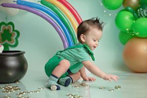 A child in green plays near a decorative rainbow, black pot and St. Patrick's Day themed balloons and decorations.