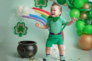 Small child in green outfit dances near St Patricks Day decorations with balloons, shamrocks and pot of gold.