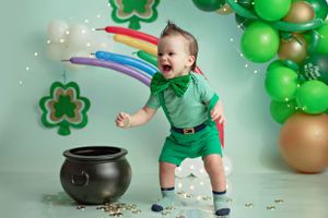 St Patrick's Day themed photo with rainbow, green balloons, shamrocks and black pot with festive decor and bubbles.