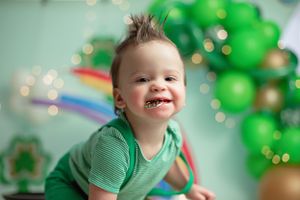 Smiling child in green shirt sits against festive backdrop with rainbow and green balloon arch decorations.