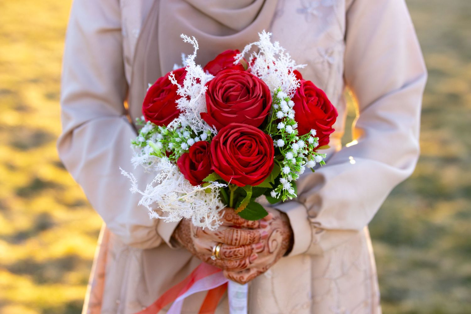 Someone in a neutral colored dress holds a romantic bouquet of red roses with baby's breath and white lace details.