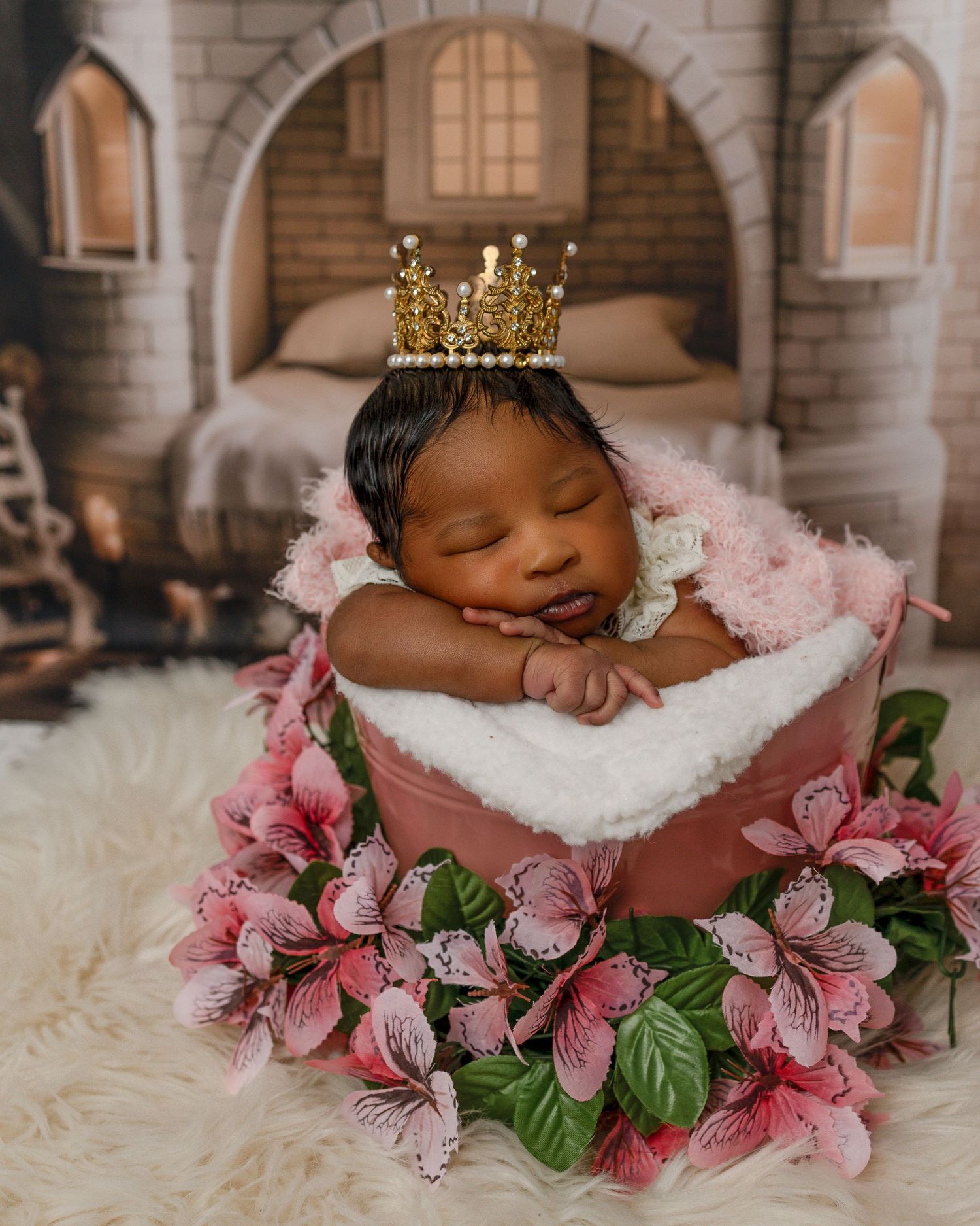 Sleeping baby in pink floral basket wearing golden crown against castle backdrop during newborn photo session.