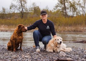 A woman sits on the riverbank with a chocolate coloured dog on her right and a gold coloured dog on her left.