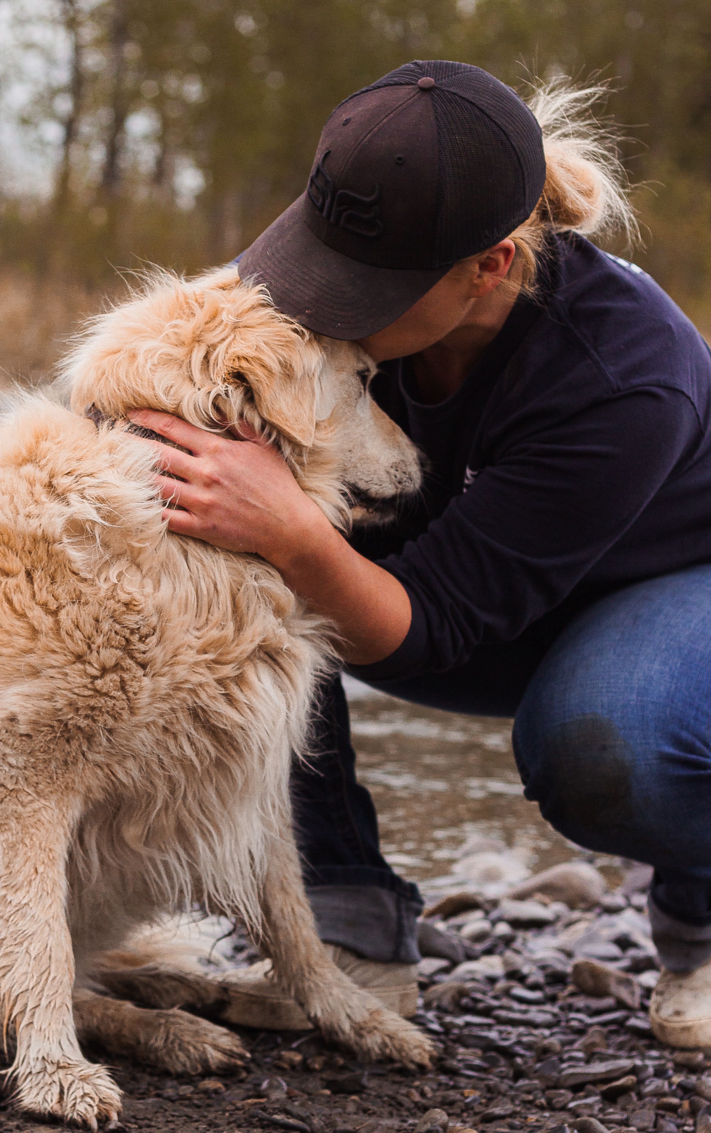 A woman hugs her golden coloured dog on a riverbank.