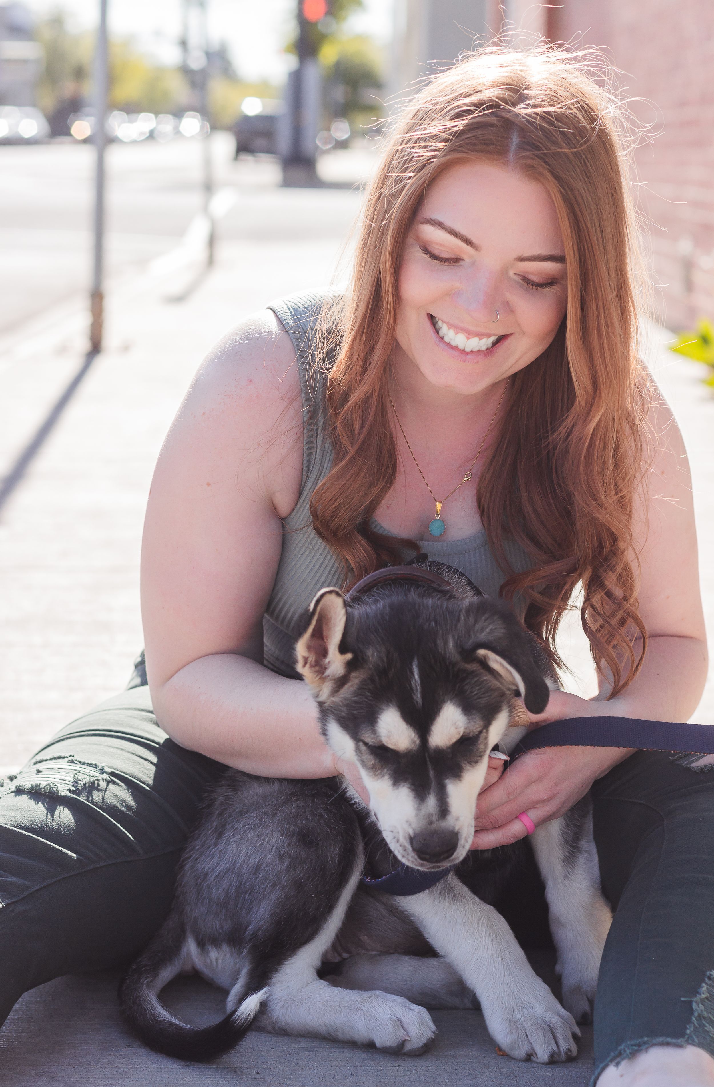 A woman sits on the sidewalk, smiling, looking down at her husky mix puppy.