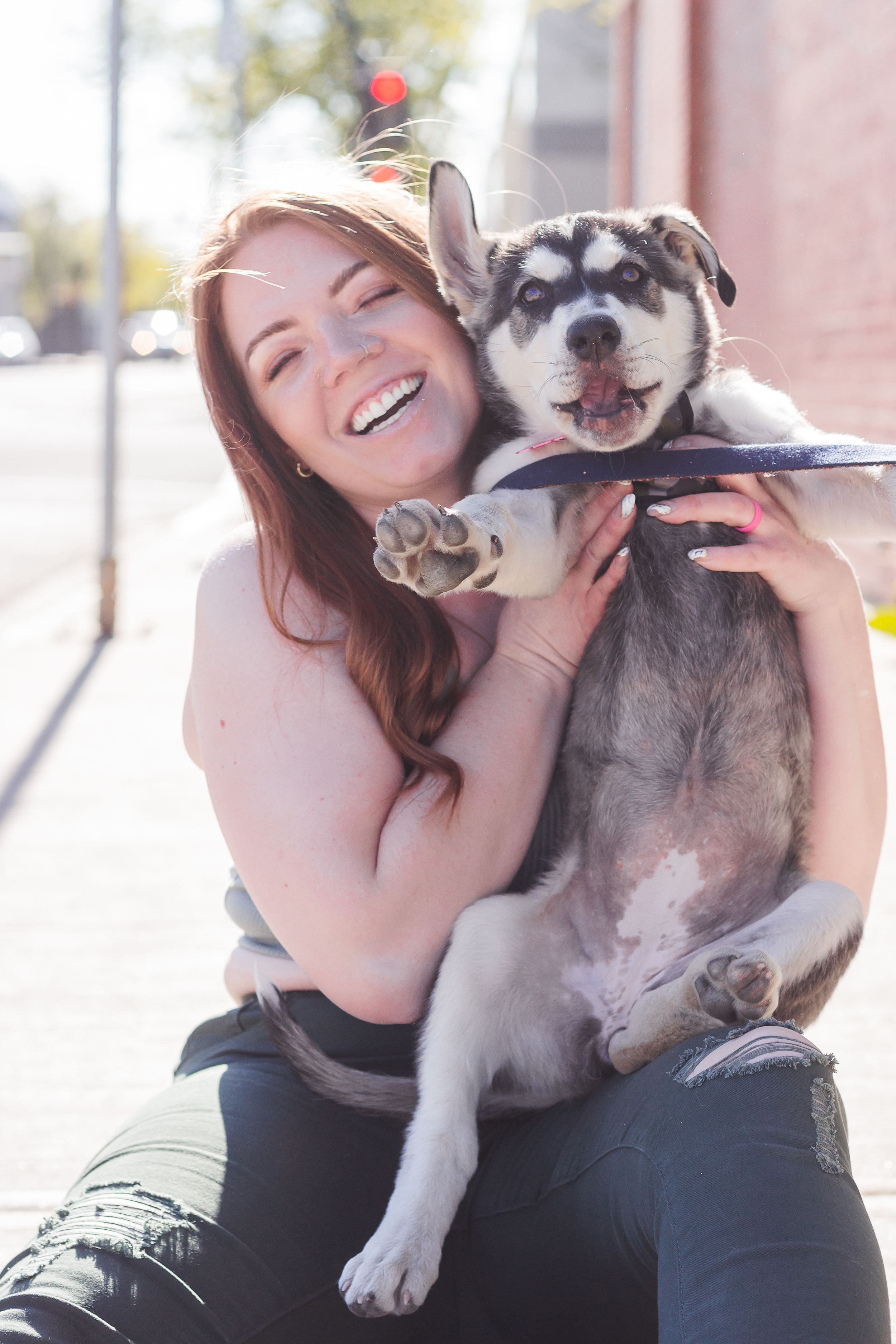 A woman sits on the sidewalk holding a husky mix puppy up.