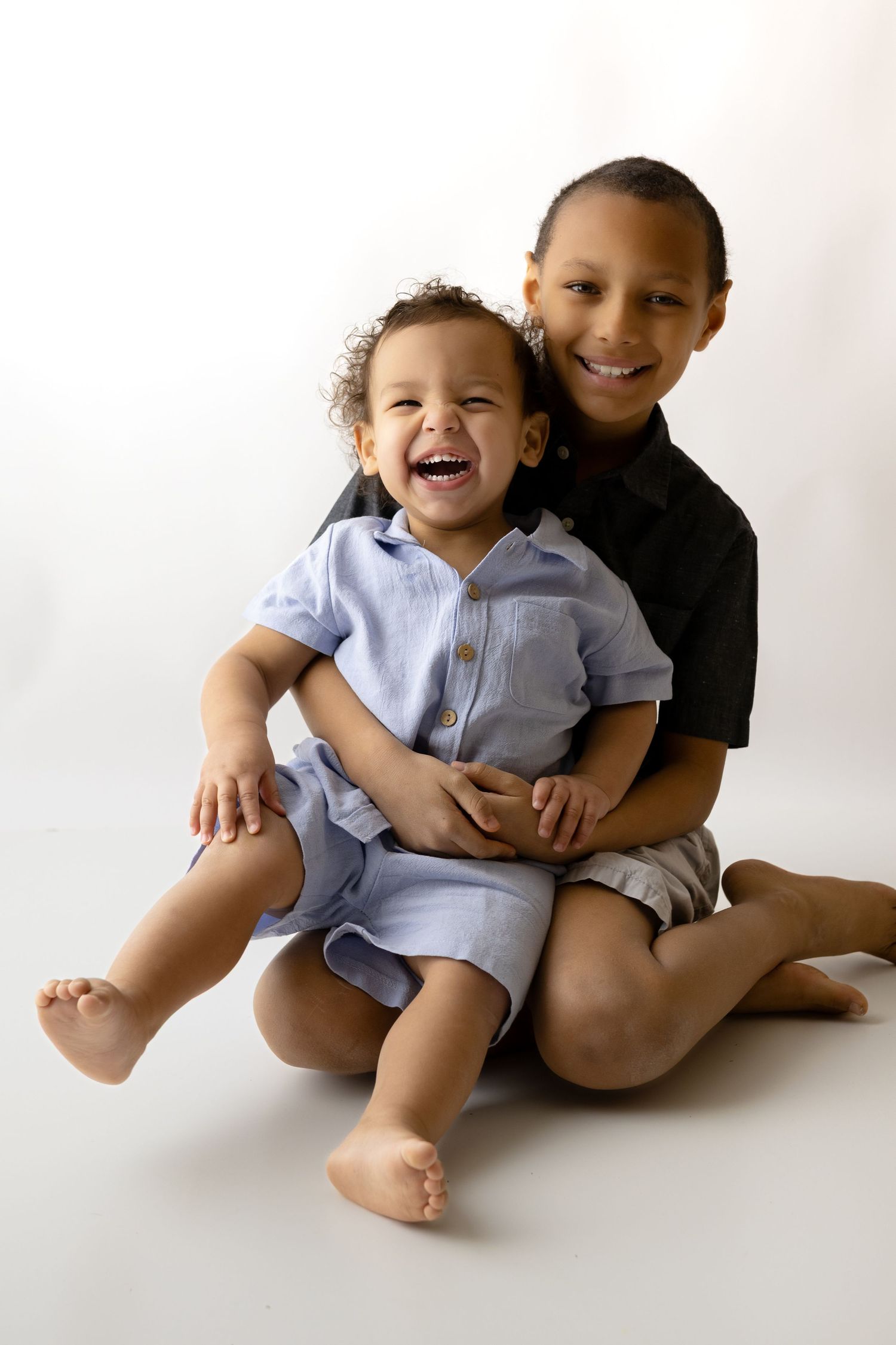 Two brothers sitting on the floor with a white backdrop, one is much older than the other. He is holding the younger and smiling/laughing