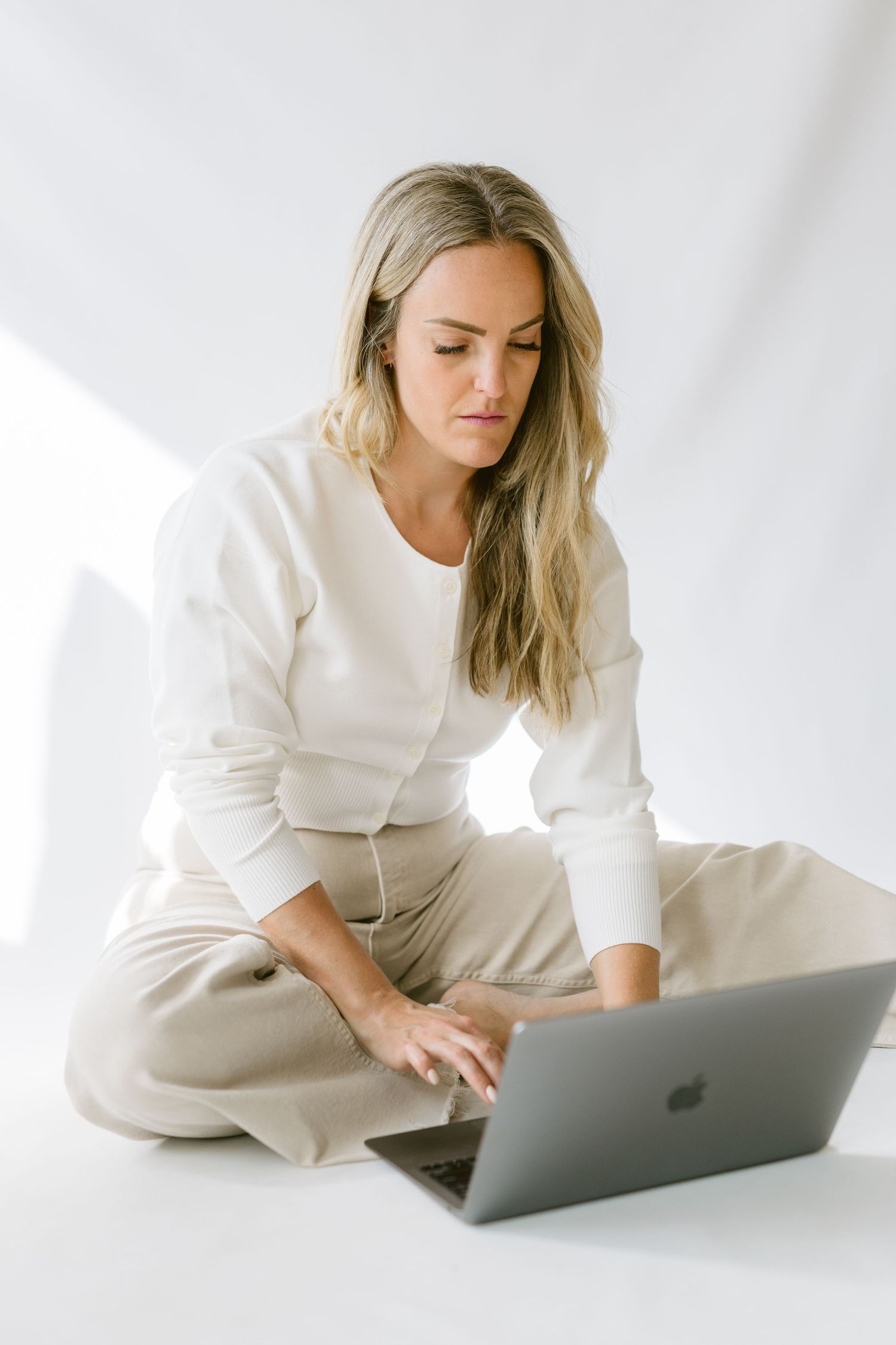 A person in white sweater and beige pants sits cross-legged on floor while working on MacBook laptop.