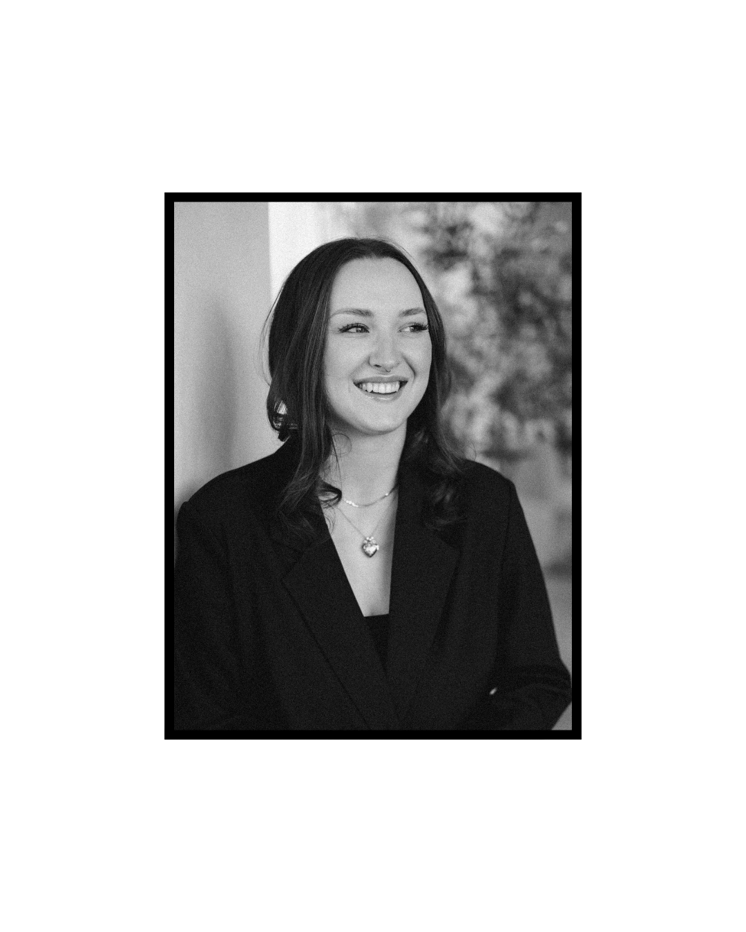 Black and white professional headshot portrait of a joyfully smiling person wearing a dark blazer against a blurred background.