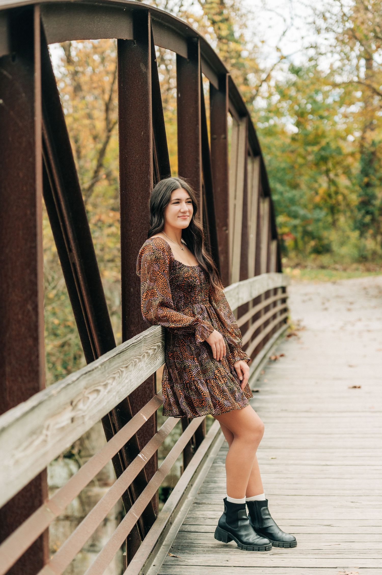 A person in a floral dress and rain boots poses on a rustic wooden bridge during autumn.