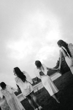 Black and white photo of four women in white dresses with angel wings standing outdoors under a cloudy sky.