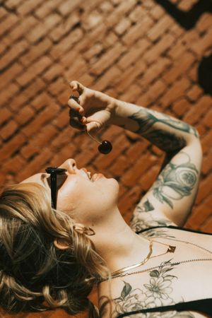A person with blonde hair and tattoos lies on brick ground holding a cherry between their fingers.