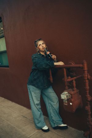 Person wearing green jacket and loose jeans poses casually against brown wall next to wooden railing.