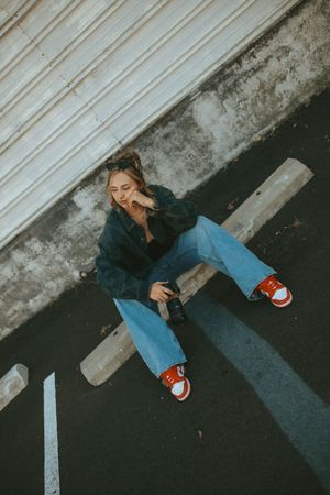 Person in blue jeans and dark jacket sitting on ground against wall wearing red and white sneakers.