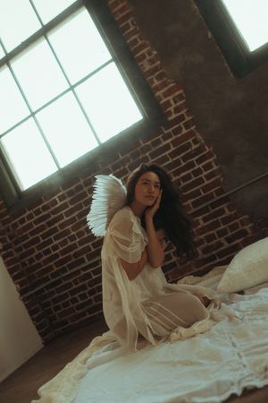 A figure in a flowing white dress sits pensively near a window in a brick-walled room with dramatic lighting.