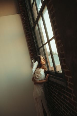 A dreamy atmospheric portrait of someone in white dress gazing pensively through a window in a brick-walled room.