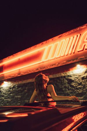 A figure rests against a car under dramatic red neon lighting at night creating a moody atmospheric scene.