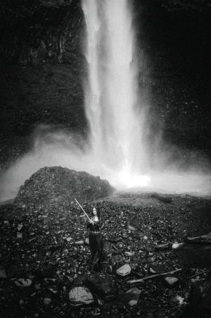 Black and white photo of a person standing before a dramatic waterfall cascading onto rocky terrain.