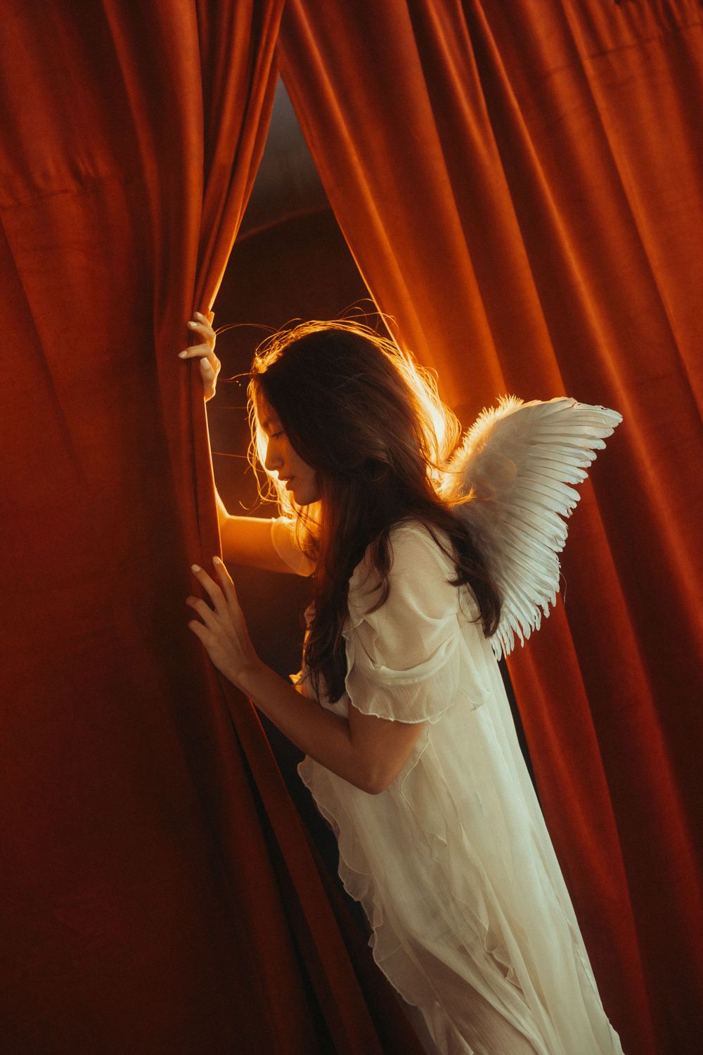 A figure in a white flowing dress and angel wings poses dramatically against red velvet curtains with fire elements.