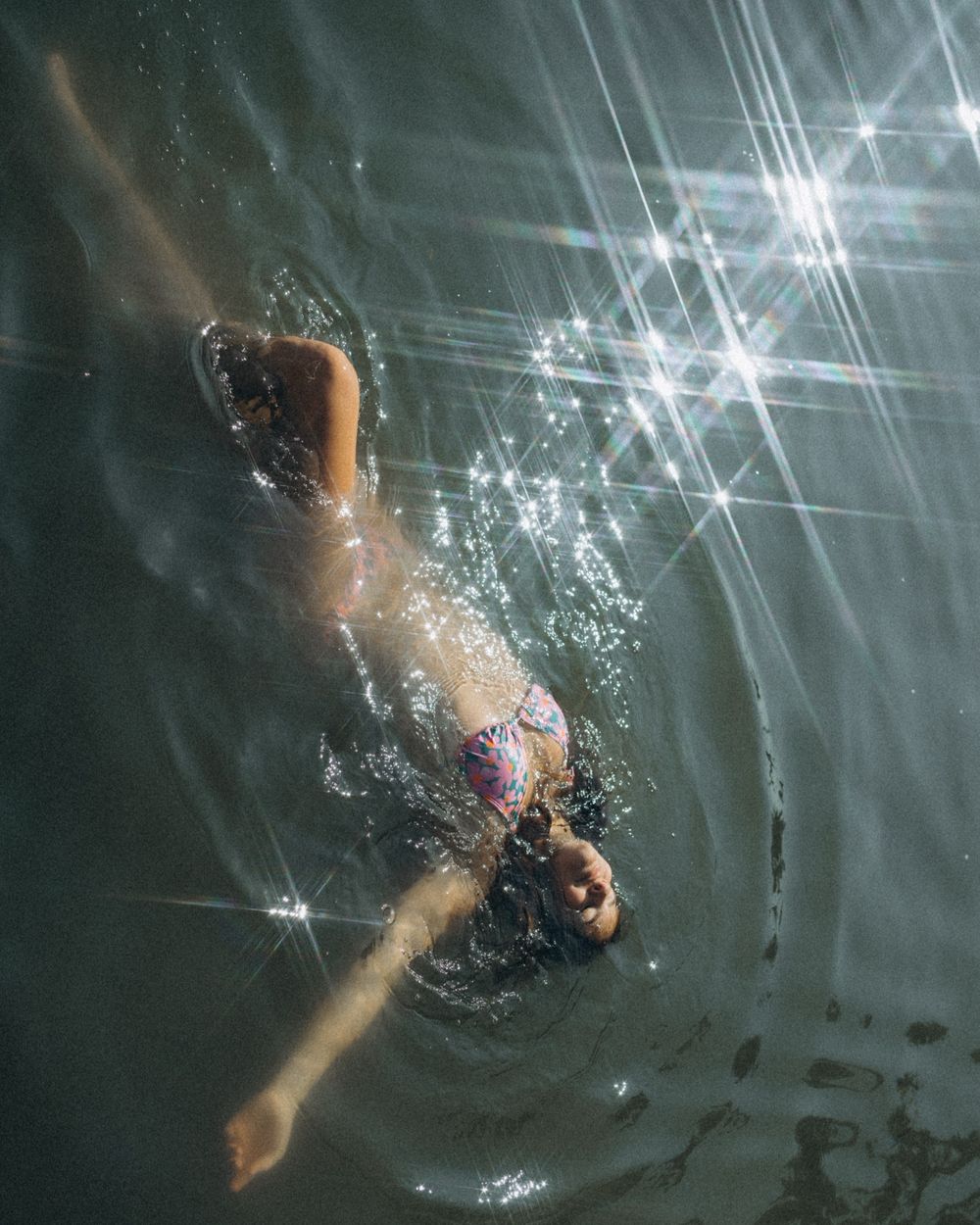 A swimmer dives underwater in a dark pool with sunlight creating sparkly beams through the water.