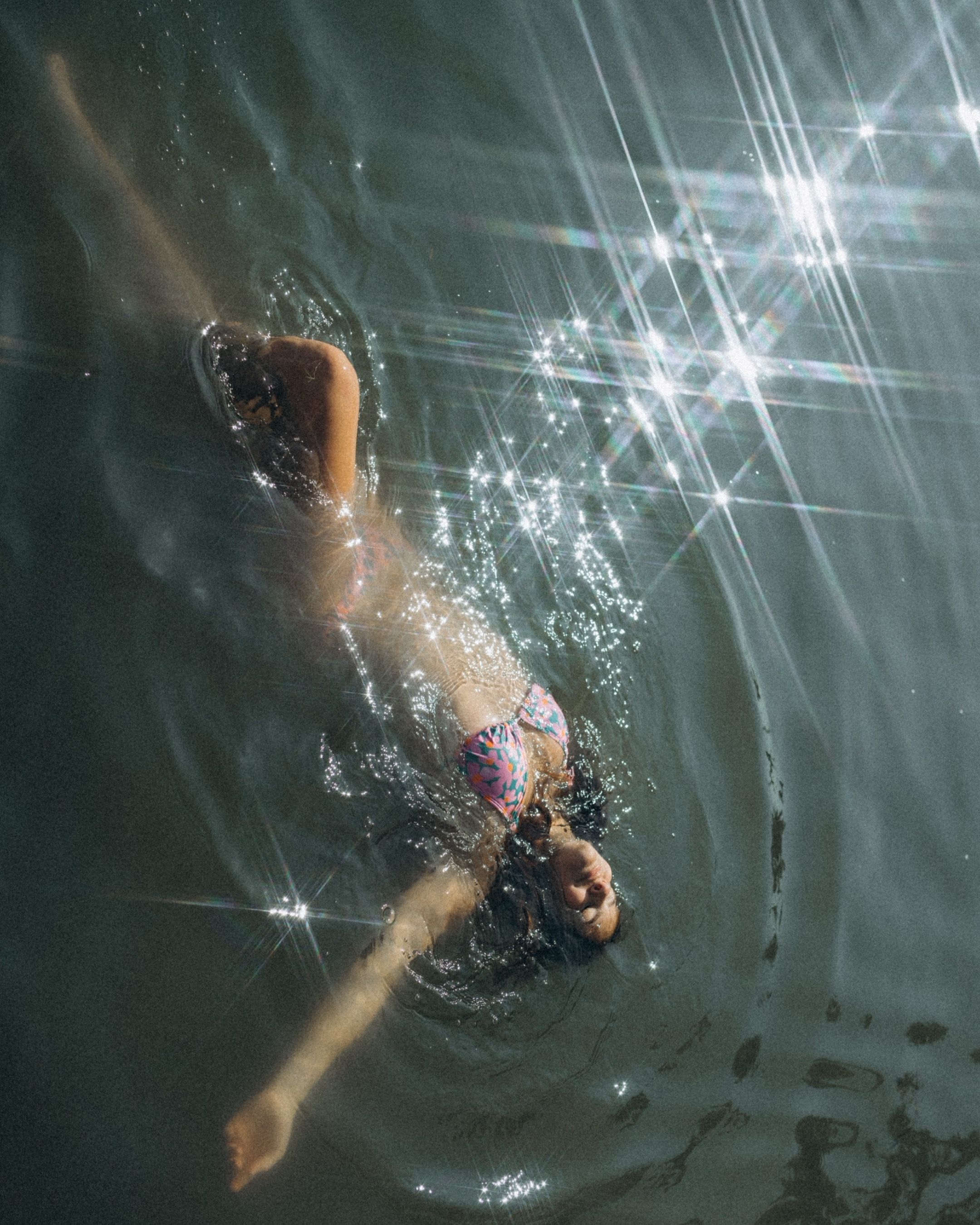 A swimmer dives underwater in a dark pool with sunlight creating sparkly beams through the water.