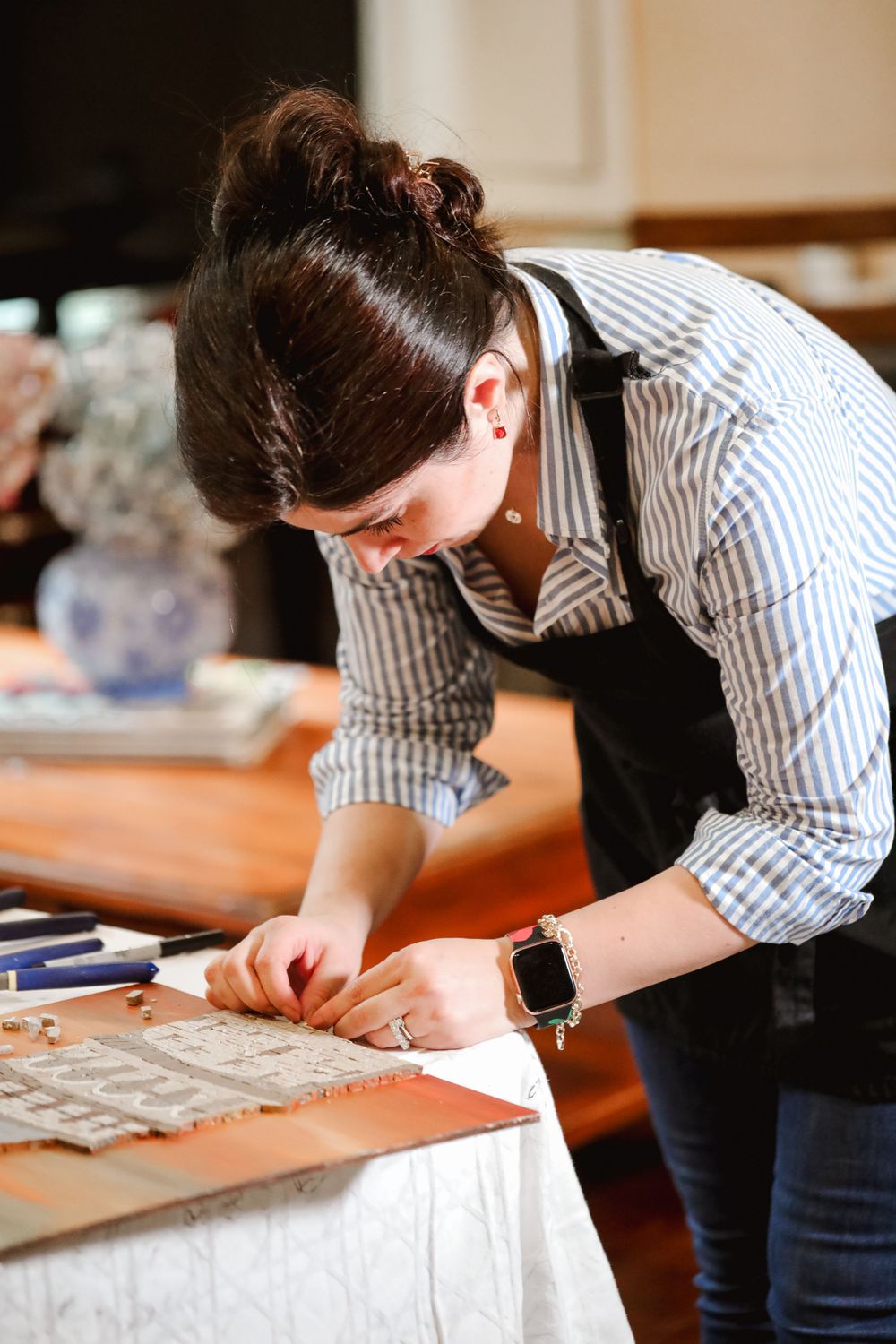 Someone wearing a striped button-down shirt and black apron works intently on a craft project at a wooden table.