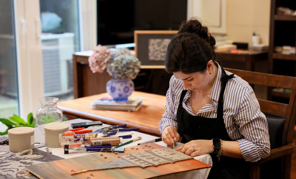 Artist carefully working on a craft project at a wooden table in a studio workspace with scattered supplies.
