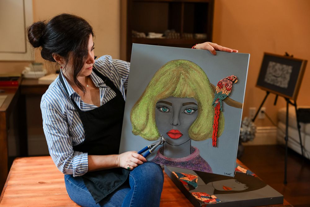 Artist in striped shirt and apron works on vibrant portrait painting of figure with neon yellow hair at wooden table.