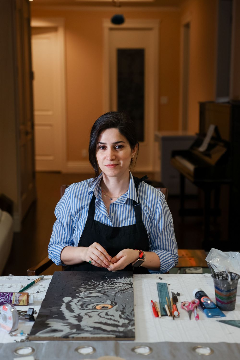 Artist in striped shirt and black apron sits at workstation with art supplies and charcoal tiger drawing in progress.