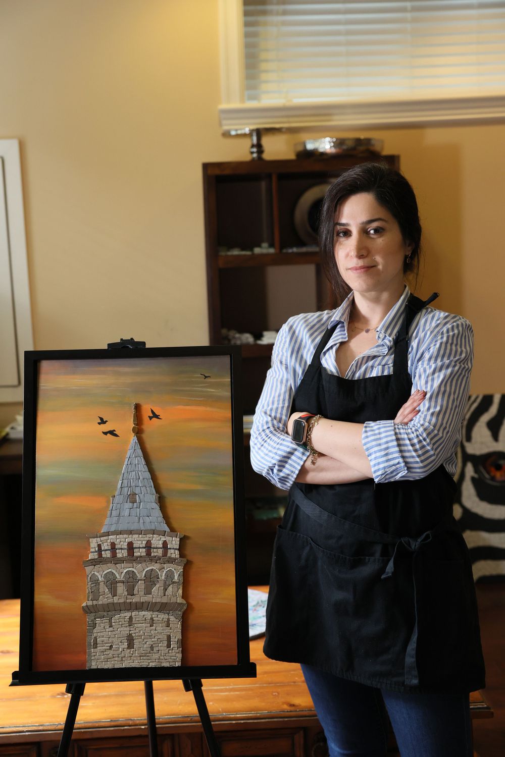 Artist in a black apron and striped shirt stands confidently next to their painting of a stone tower against a sunset sky.