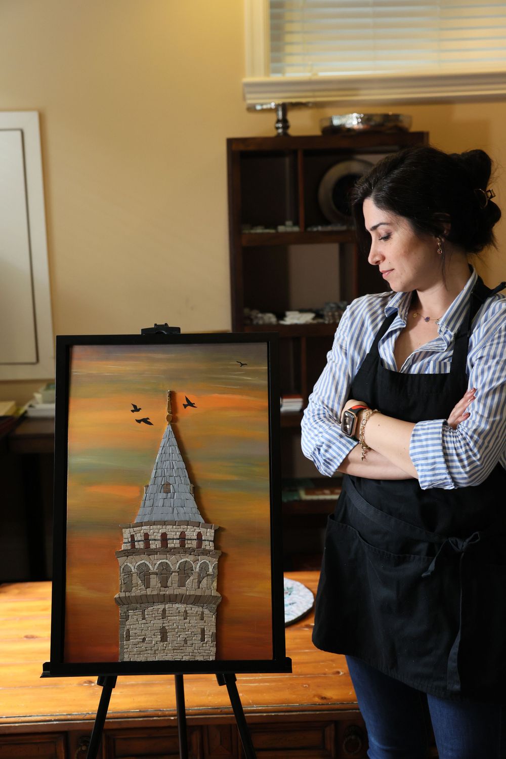 Artist in striped shirt and black apron stands beside easel displaying painting of tower against orange sunset sky.
