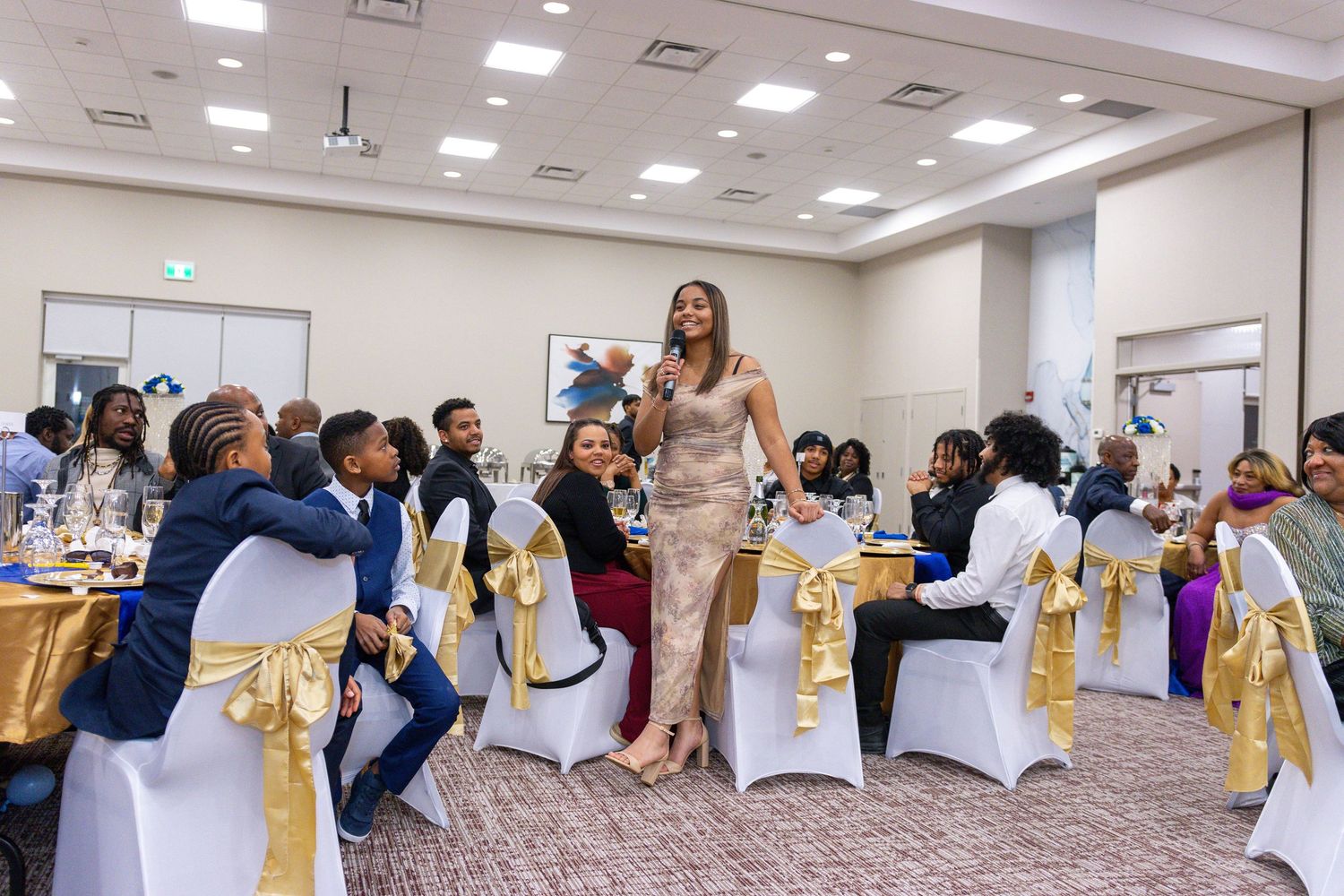 Guests in formal attire gather at decorated tables with white chair covers and gold accents during an elegant event celebration.