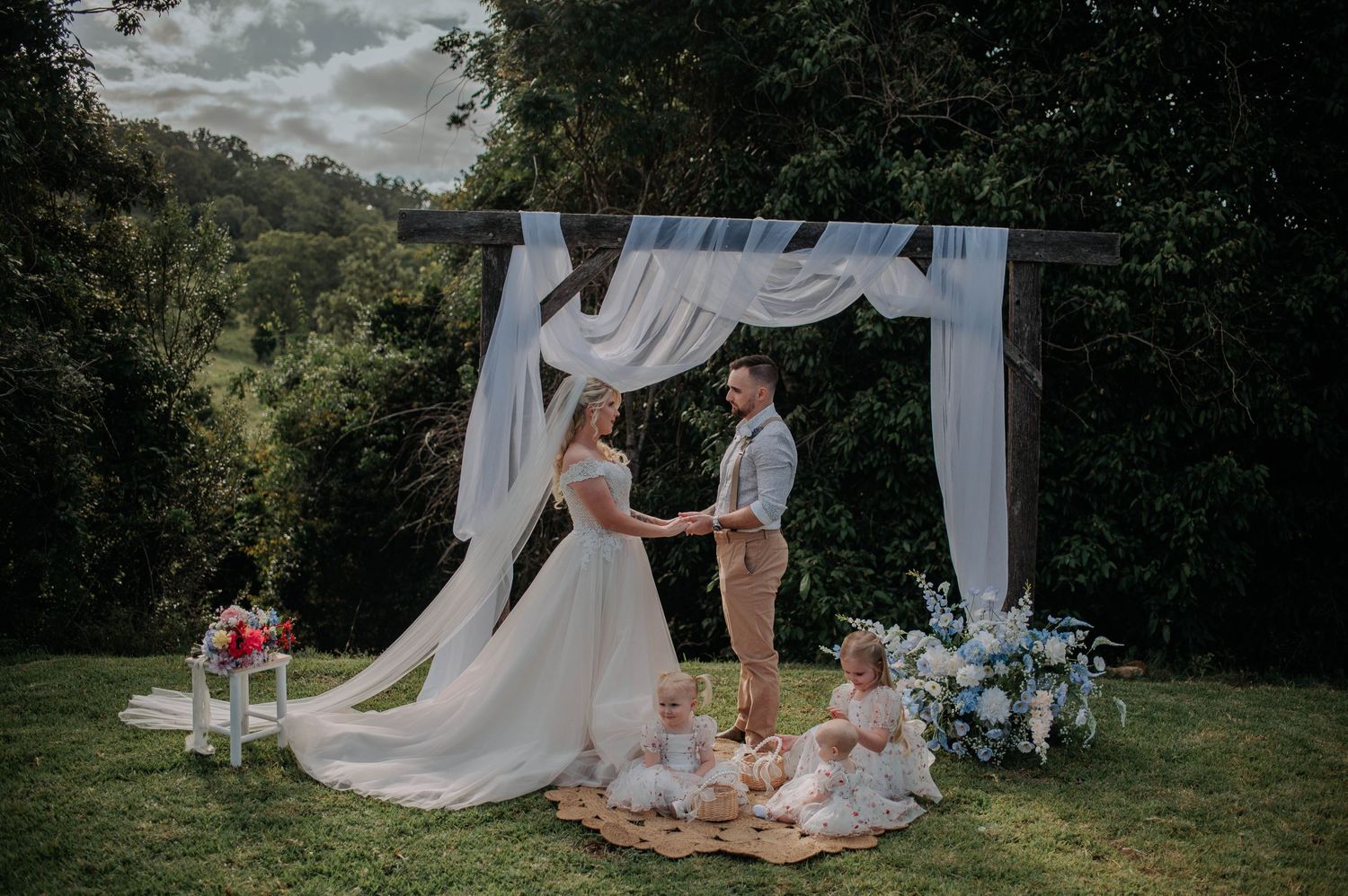 A couple exchanges vows under a white draped arch decorated with flowers.