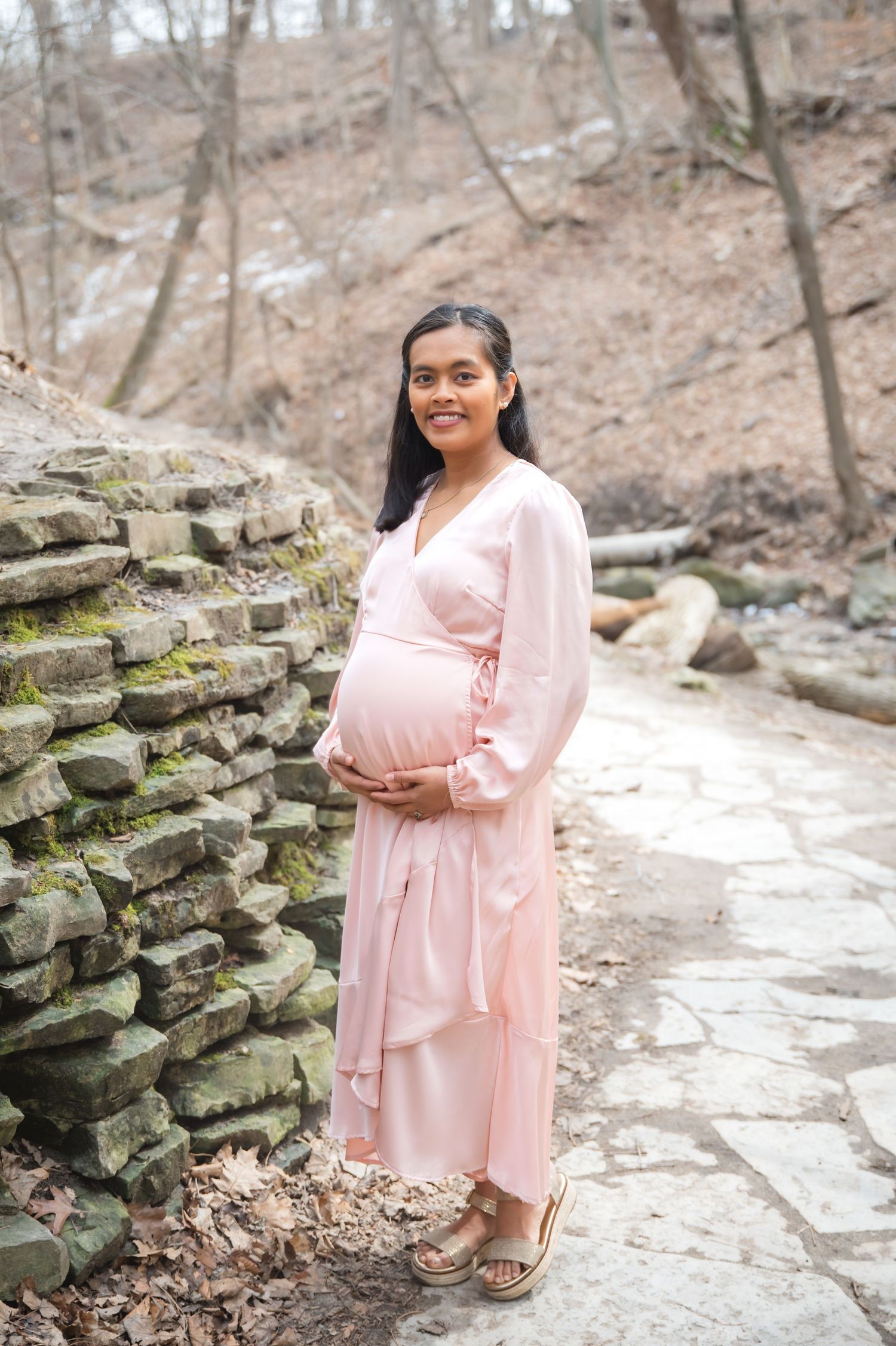A person in a flowing pink dress stands next to a stone wall on a woodland path during winter.