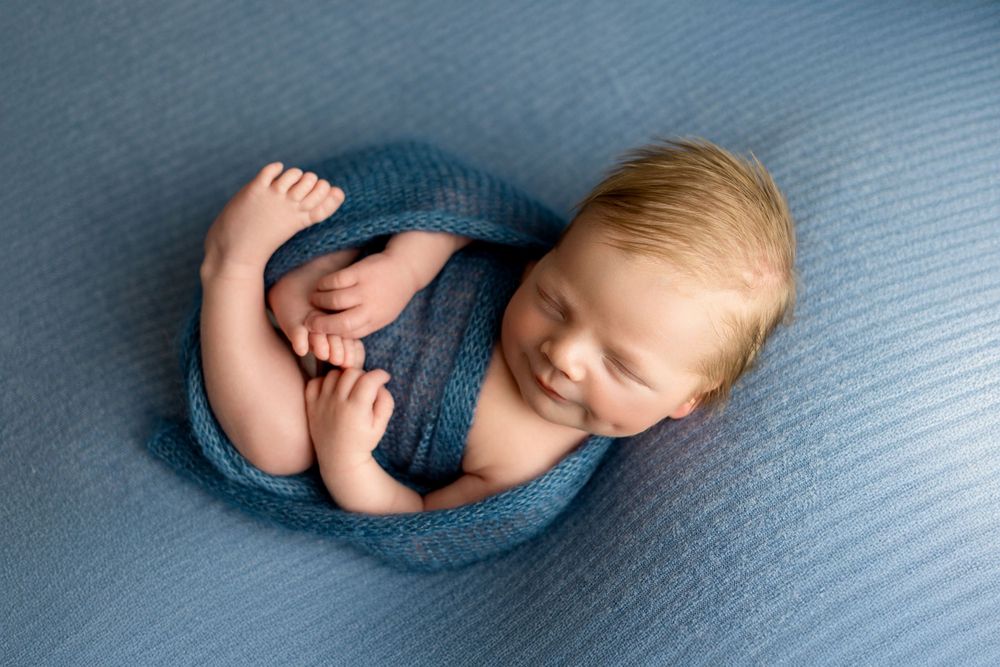Sleeping newborn baby curled up in cozy blue wrap on textured blue background during professional photo session.