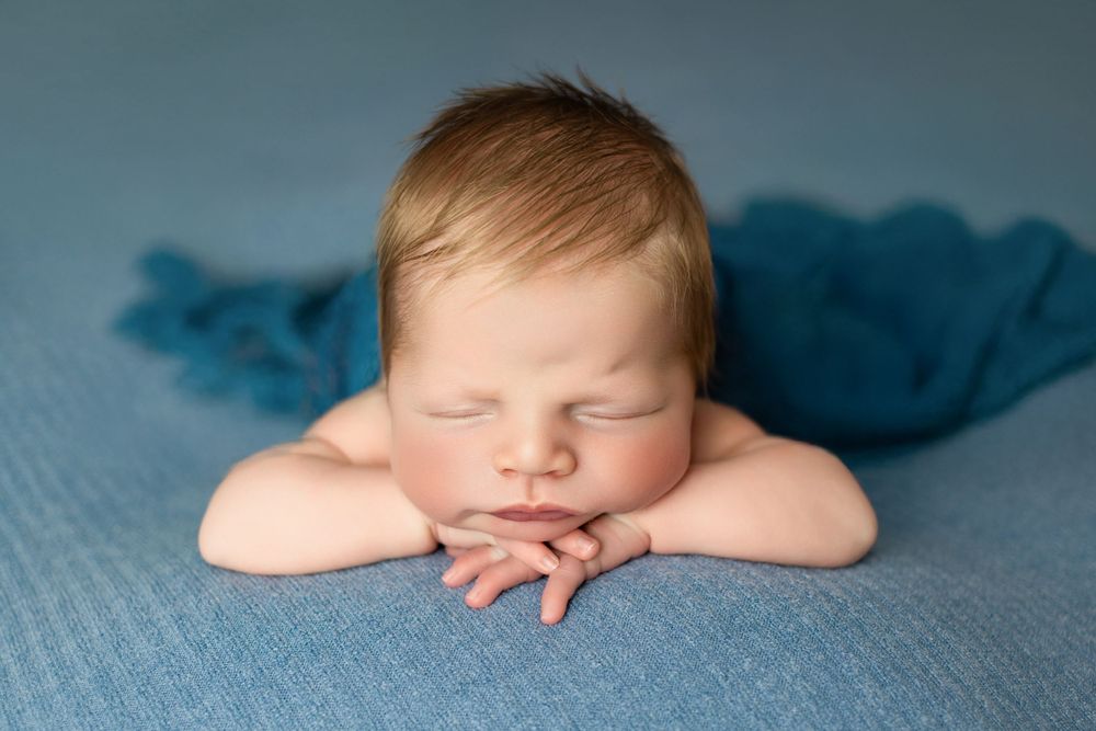 Peaceful sleeping newborn rests with folded arms on soft blue blanket during professional photography session.