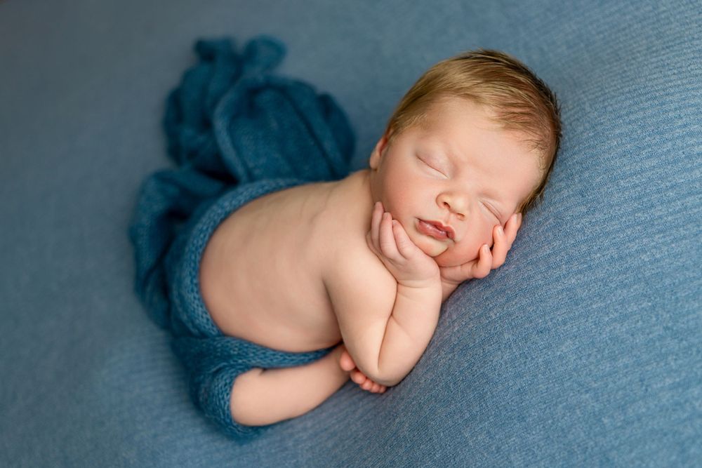 Sleeping newborn baby wrapped in teal fabric against blue background in studio portrait photography.