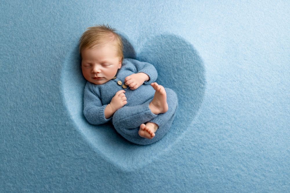 Sleeping baby in blue clothing nestled in heart-shaped plush fabric during newborn photo session.