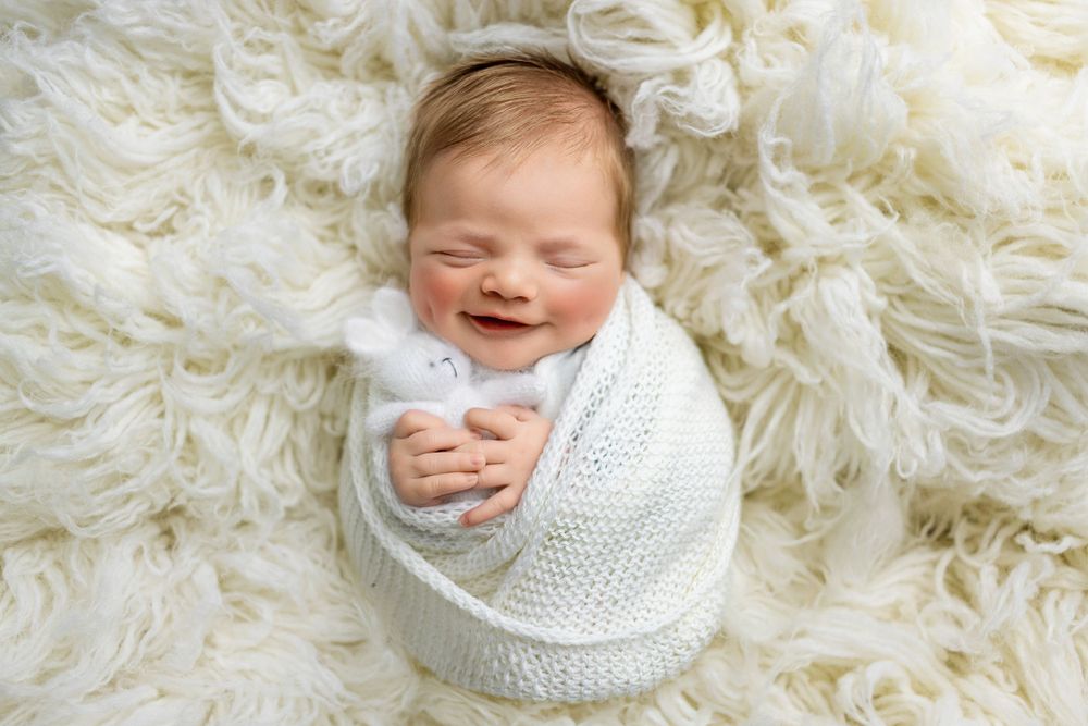 A newborn baby wrapped in a white knitted blanket smiles while lying on a soft cream-colored fur background.