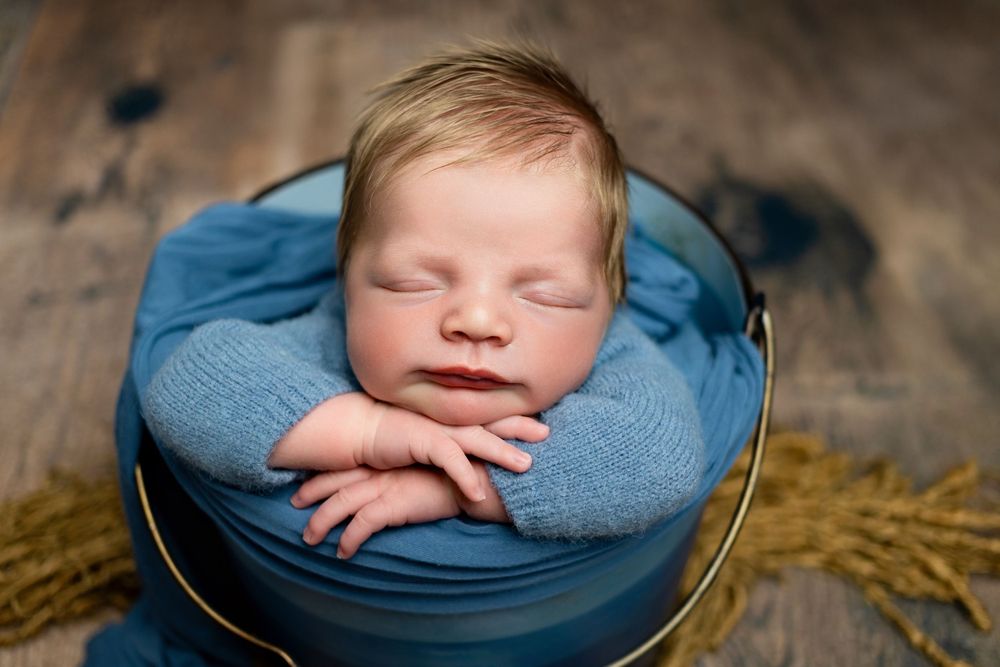 Sleeping newborn in blue wrap nestled in rustic bowl with hay accents.