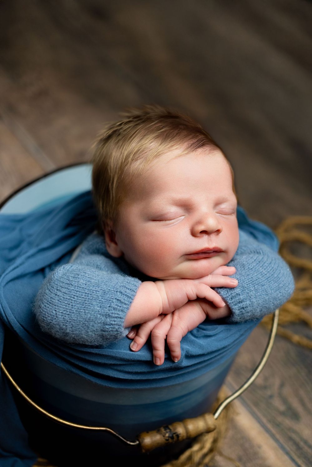 Newborn baby sleeping peacefully in blue blanket inside vintage wooden basket on hardwood floor.