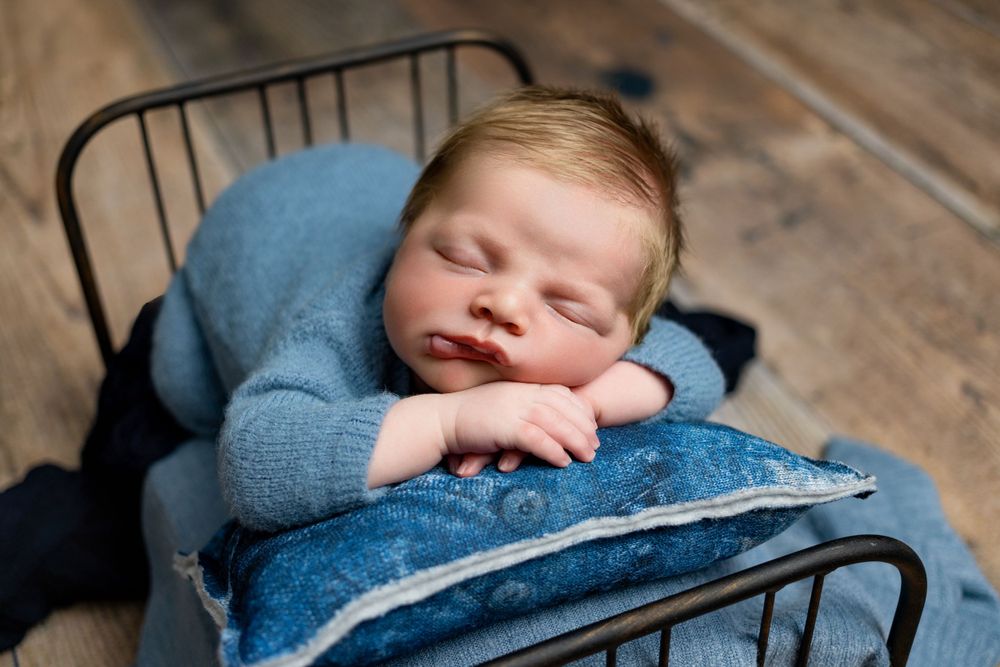 A sleeping newborn in a blue sweater rests peacefully on a dark metal bed frame with wooden floors beneath.