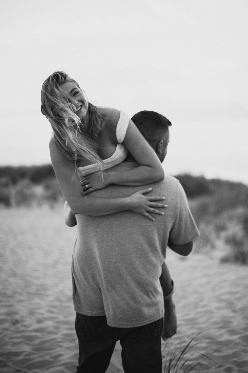 A romantic black and white photo shows a couple embracing on a sandy beach at sunset.