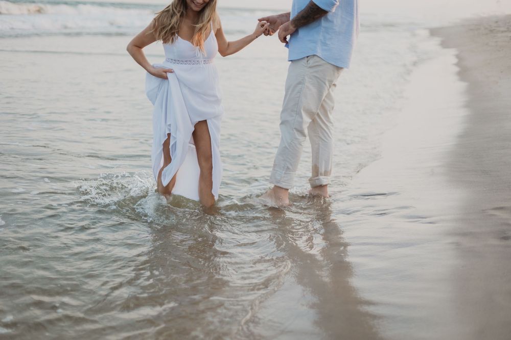 A couple in white and blue casual attire hold hands while walking in shallow ocean waves on a beach.