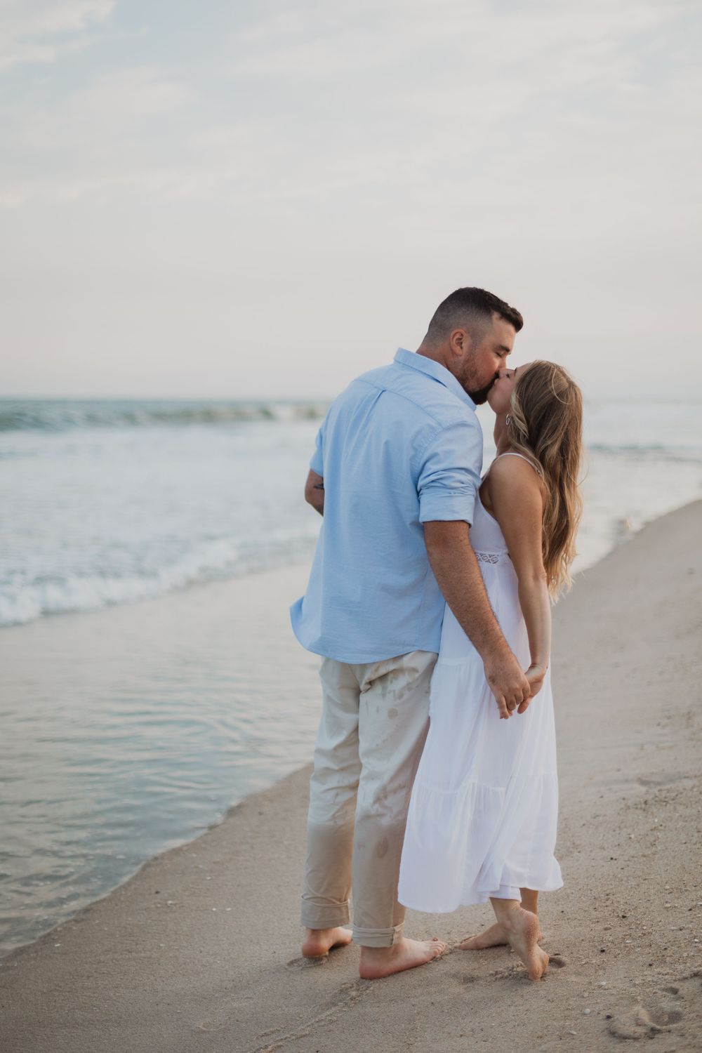A couple shares a romantic moment while standing barefoot on a beach at sunset with gentle waves in the background.