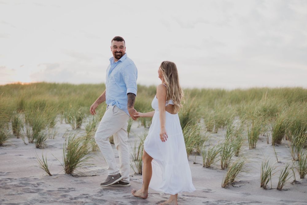 A couple walks hand in hand through beach grass and sand dunes during a sunny summer evening.