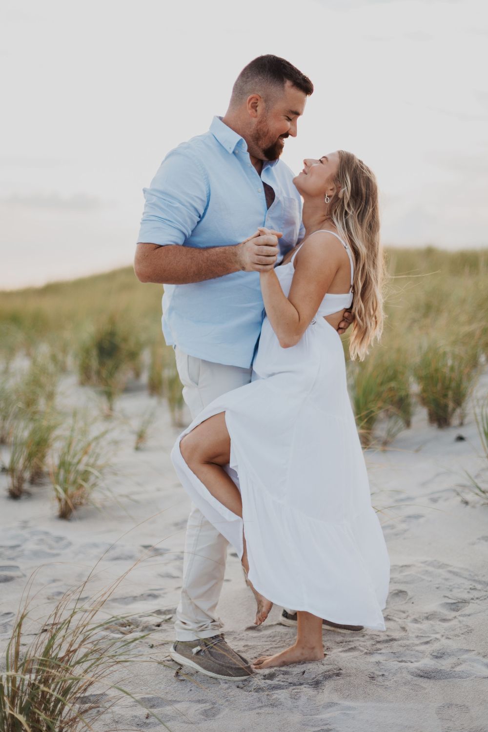 A romantic couple embraces on a sandy beach with dunes and beach grass in the background at sunset.