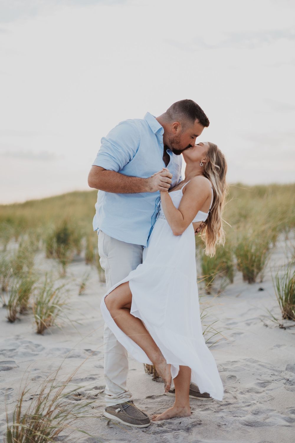 A couple in light blue and white clothing dance together on a sandy beach with grass-covered dunes.