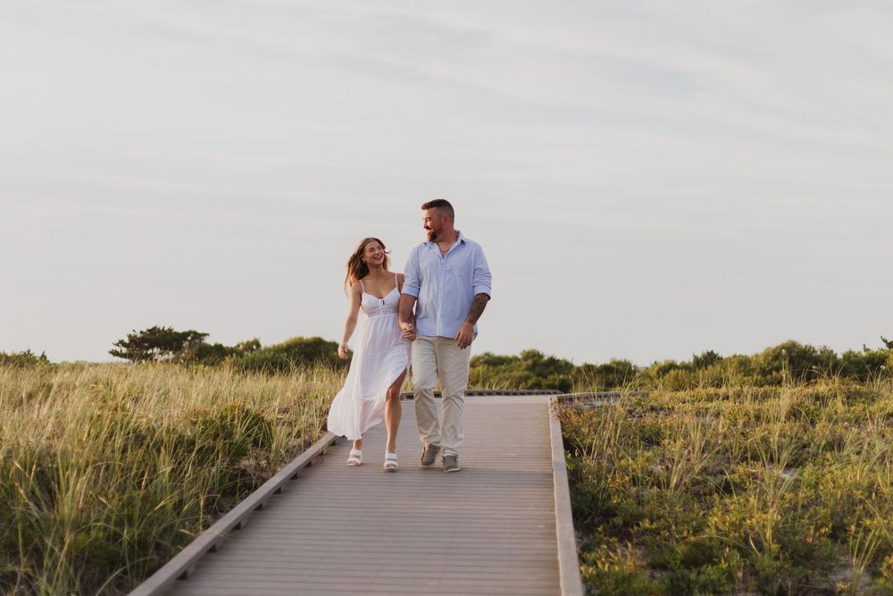 Two people walk hand in hand along a wooden boardwalk through grassy dunes at sunset.